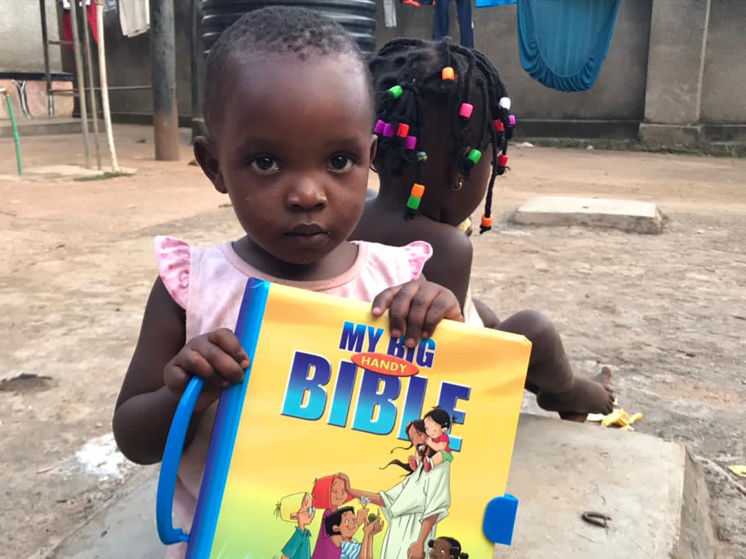 A young girl sits on the ground holding a yellow "My Big Bible" picture book.