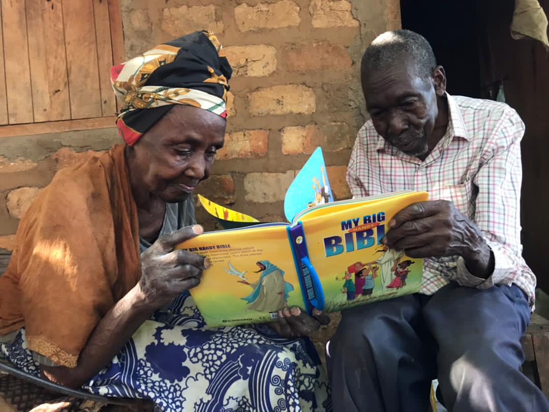 An elderly couple sits together outdoors reading a new yellow "My Big Bible" picture book.