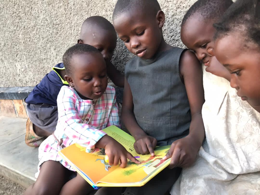 Three children sit together outdoors reading a yellow "My Big Bible" picture book.