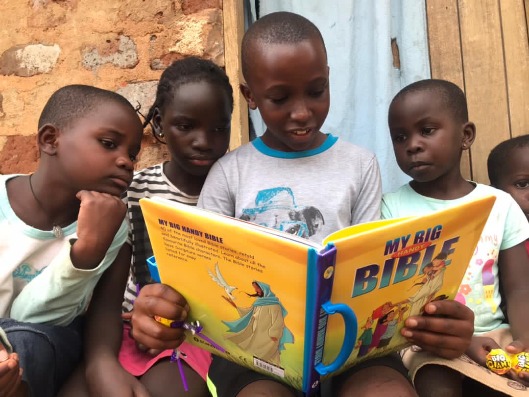Two children and a baby read an open "My Big Bible" picture book together against a brick wall.