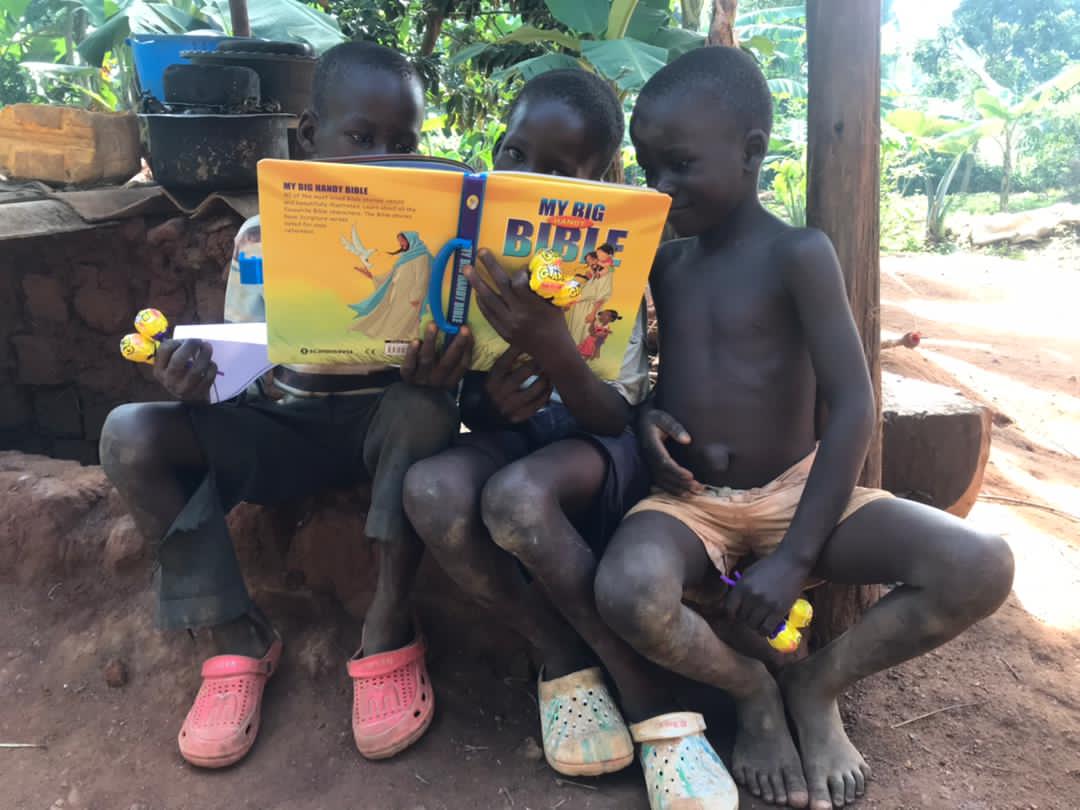 Two children sit on the ground outdoors reading an open "My Big Bible" picture book, sandals scattered nearby.