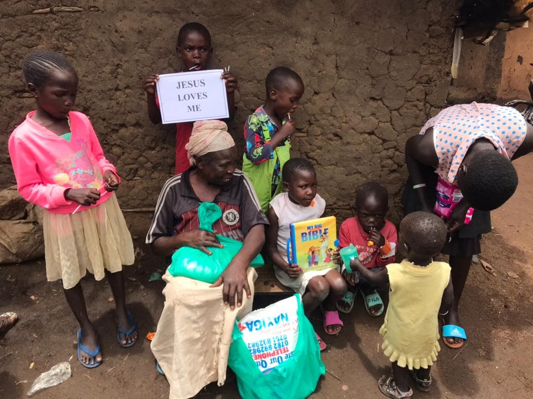 A woman and group of children stand outdoors near a mud wall, a child holding a "JESUS LOVES ME" sign.