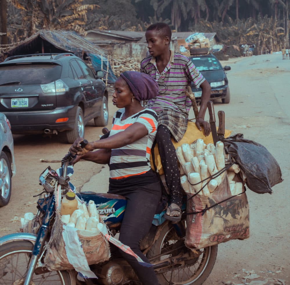 A woman rides a motorcycle loaded with sacks of cassava roots while a man walks alongside on a street.