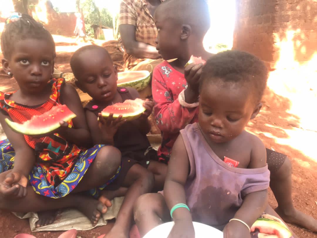 A group of young children sit outdoors on red dirt eating slices of watermelon.