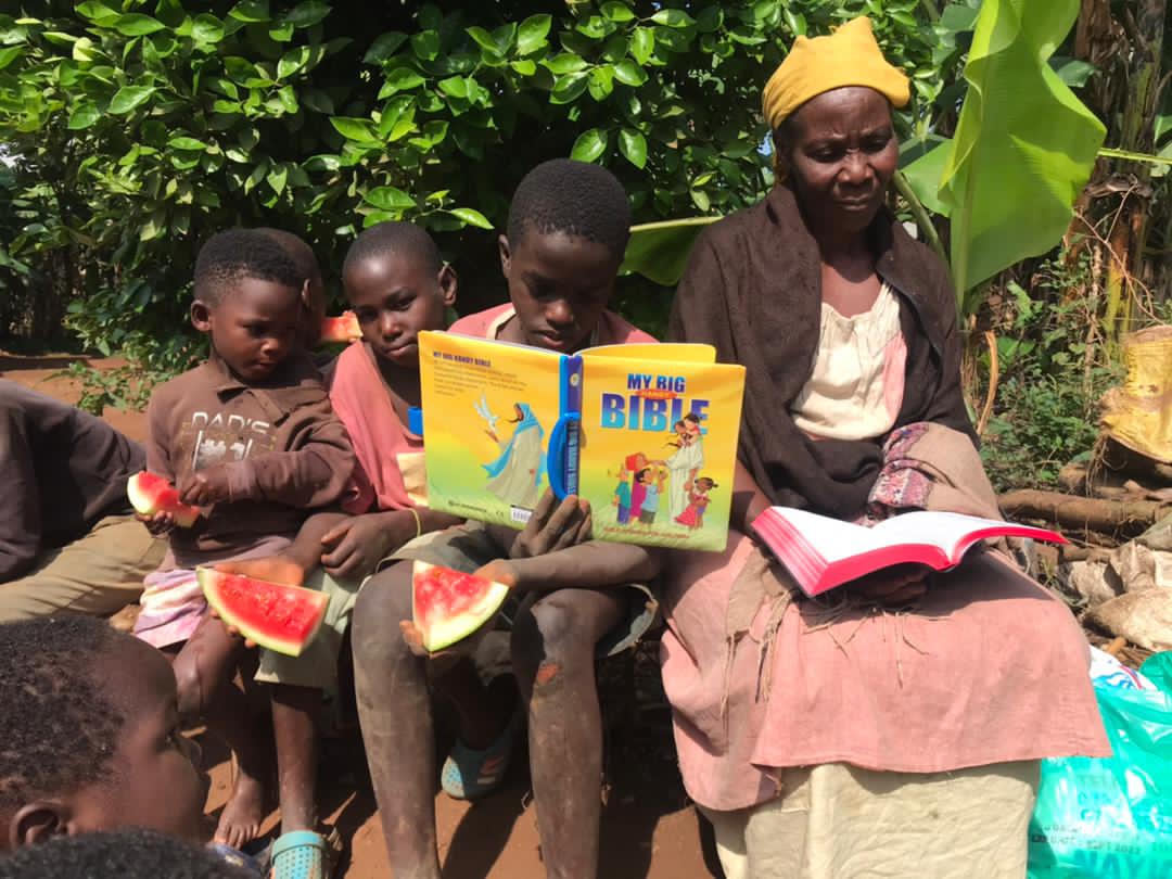 An elderly woman sits with two children, one eating watermelon and the other holding a My Big Bible board book.