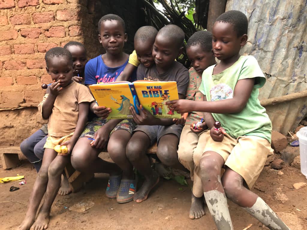 A group of children sit together on a bench, one holding a My Bible board book.