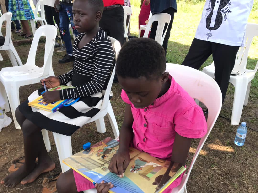 A boy in a pink shirt sits on a white plastic chair outdoors reading a picture book at Sunday school.