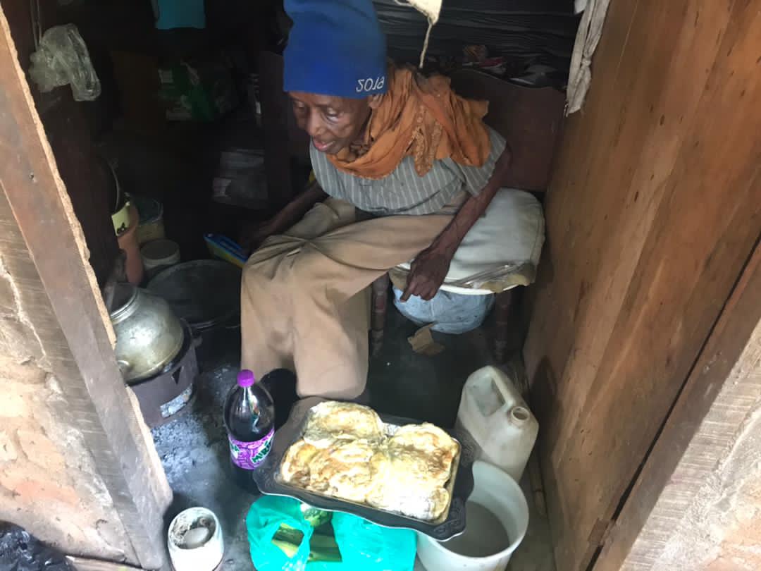 An elderly woman sits on a stool beside a wooden crate structure with food items nearby.