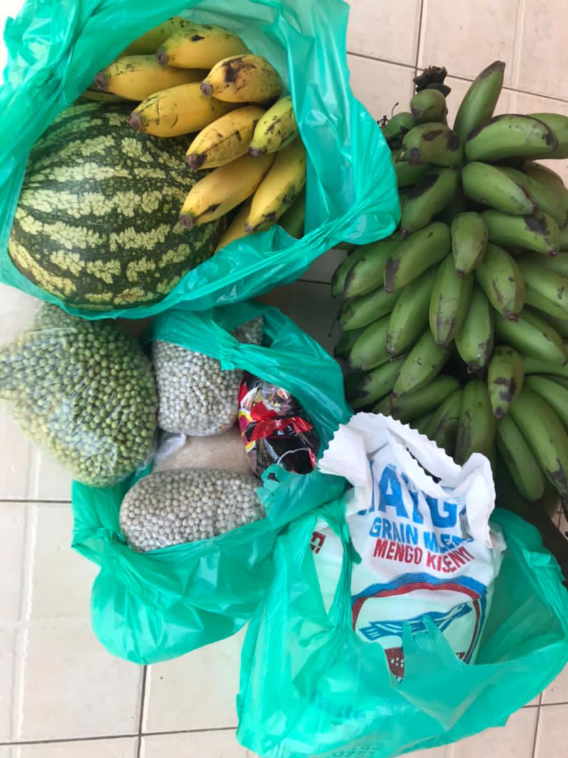 Green plastic bags filled with watermelon, bananas, and other produce sit on a tile floor.