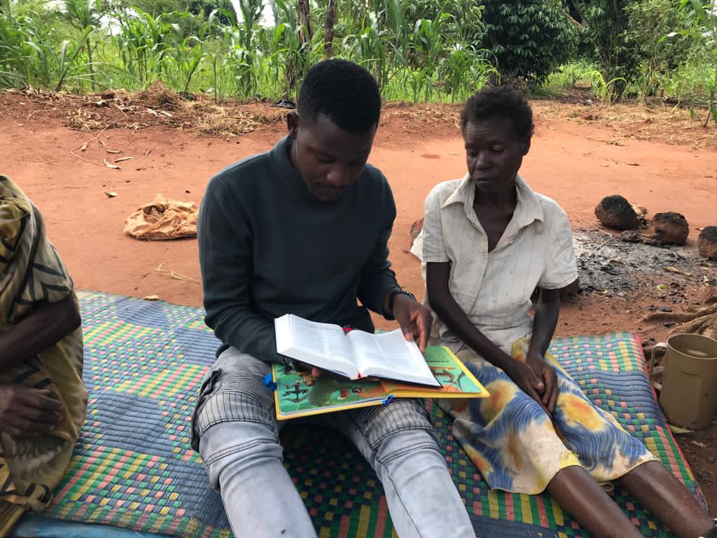 Two children sit together outdoors on a mat reading a picture book in rural Uganda.