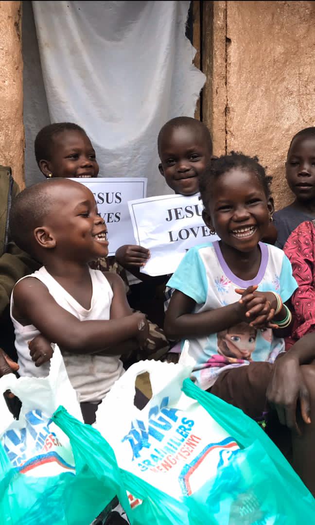 A group of children sit together outdoors smiling, holding a green plastic bag of food supplies.