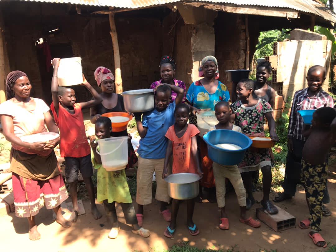 A woman and several children stand under a thatched shelter holding basins of white food such as posho.