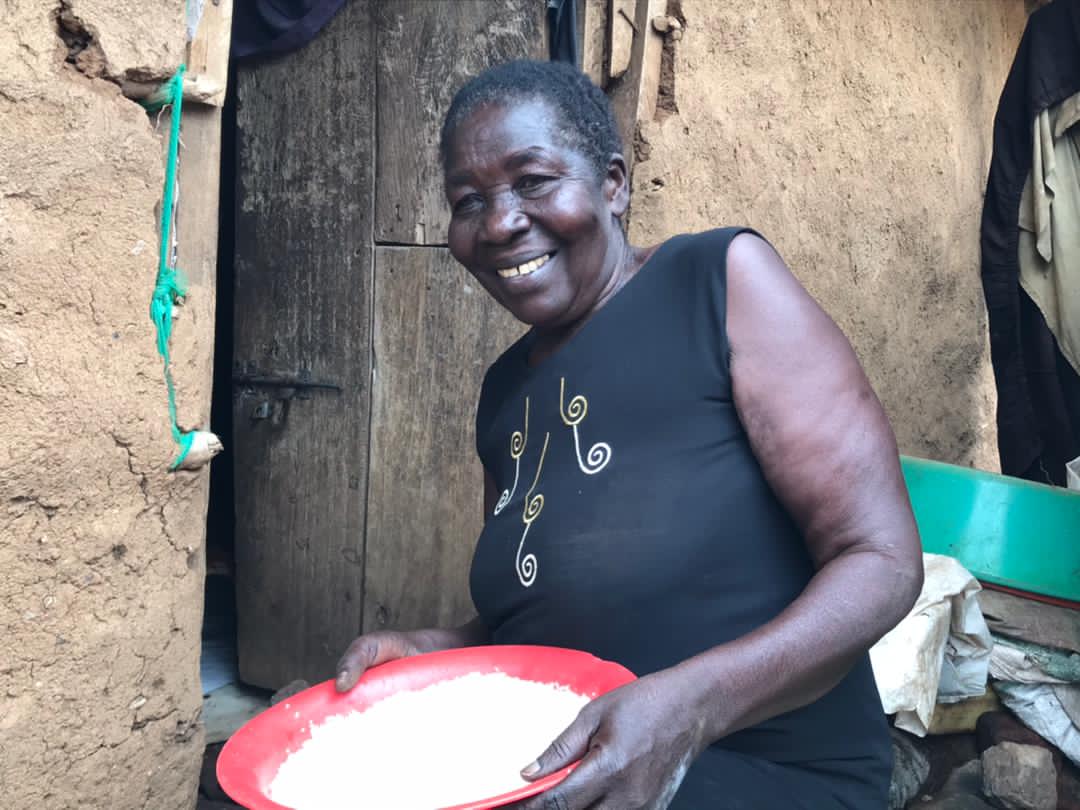 A smiling elderly woman holds a pink plate of food outside a mud home.