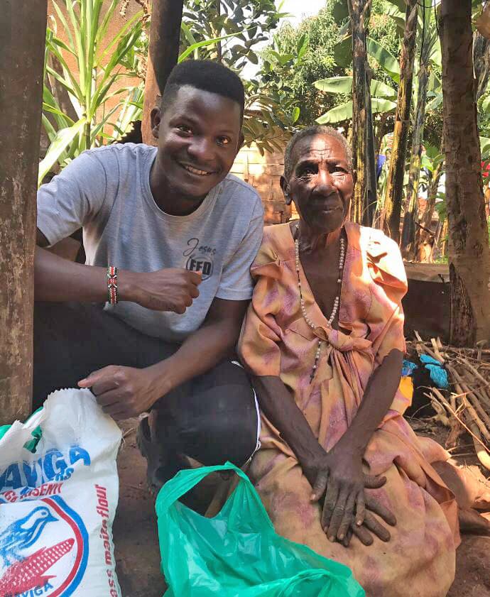 A young man crouches beside an elderly woman with a green bag of food supplies at their feet.