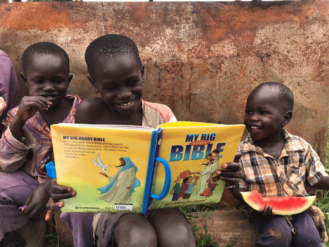 Two children hold an open "My Big Bible" picture book while a younger child eats watermelon, rust-colored wall behind them.