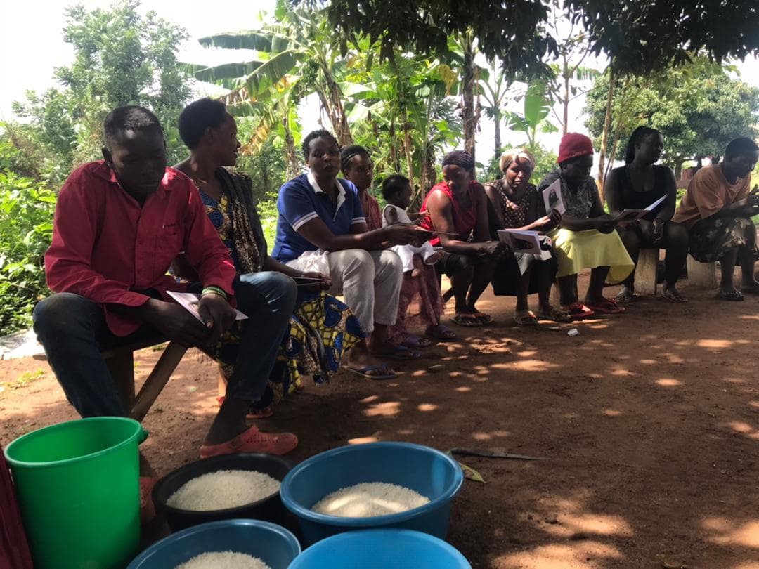 Group of women and children seated in a circle outdoors with buckets and basins of food