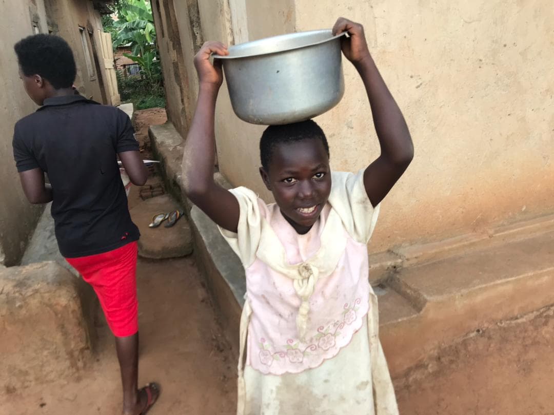 Girl in a light dress carrying a metal basin of food on her head near a brick building