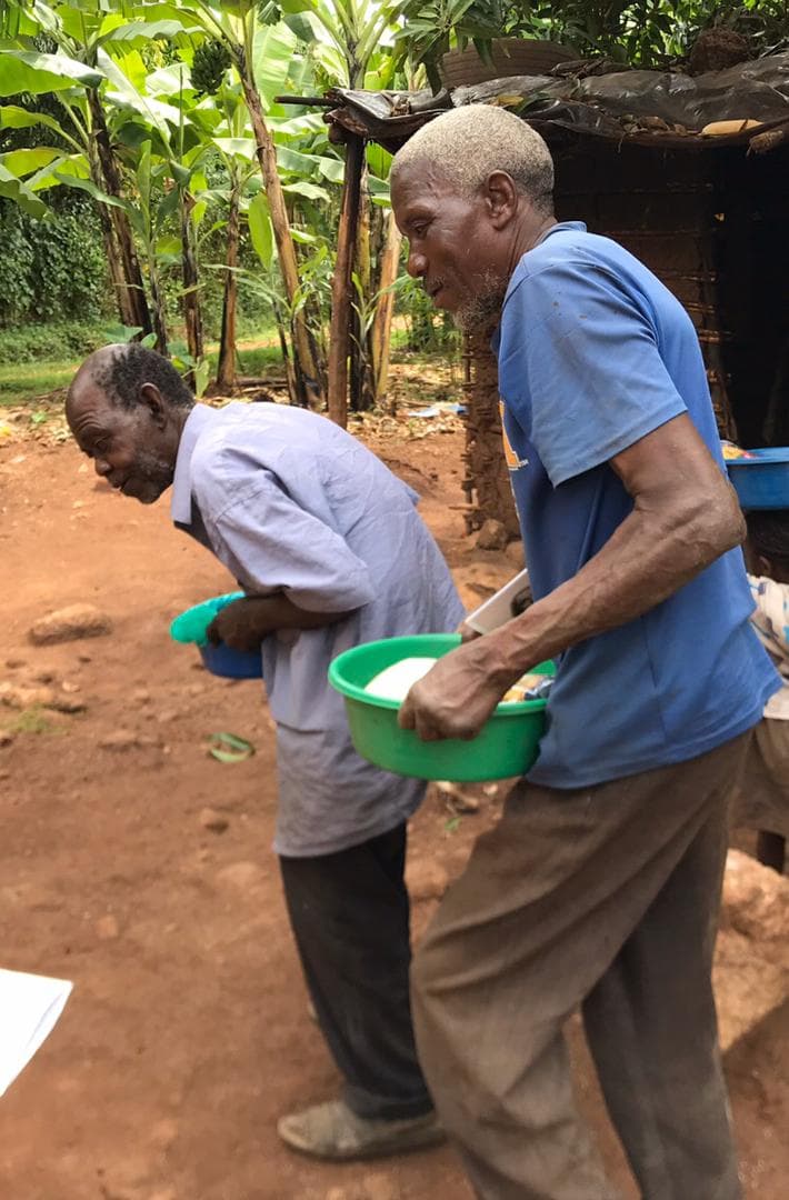 Two elderly men carrying green basins of food outside a thatched-roof home