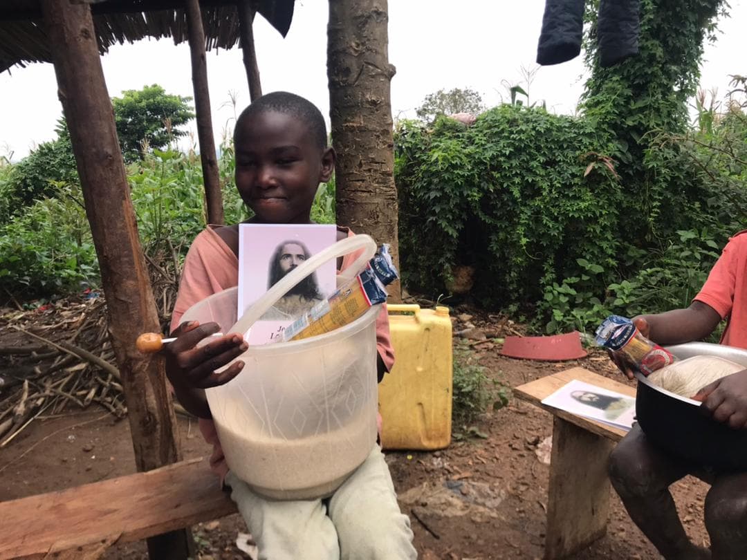 Boy holding a gospel tract and a basin of food beside a jerrycan under a thatched shelter