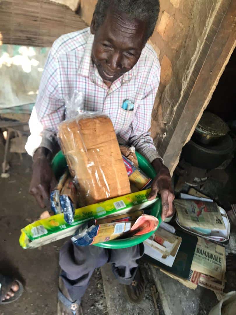 An elderly man in a checkered shirt holds a basin filled with bread, drinks, and food items indoors.