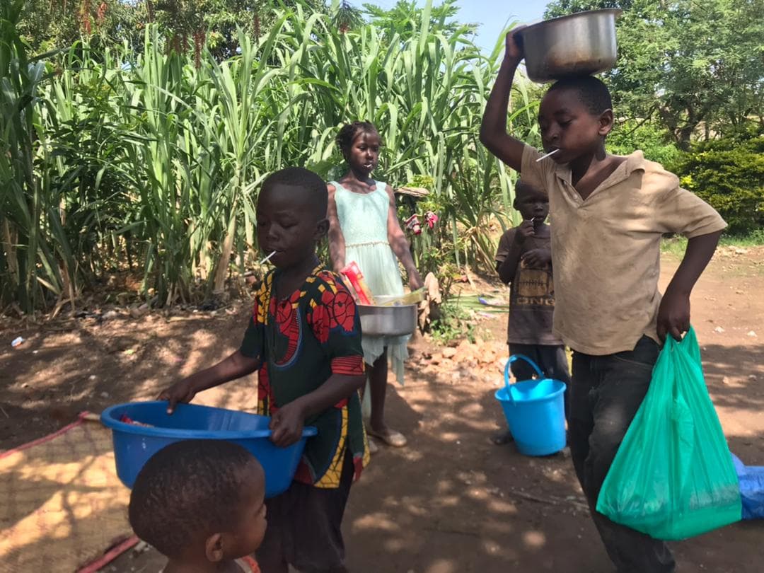 Children stand outdoors near a sugarcane field, one balancing a bowl on her head, holding a blue basin and green bag.