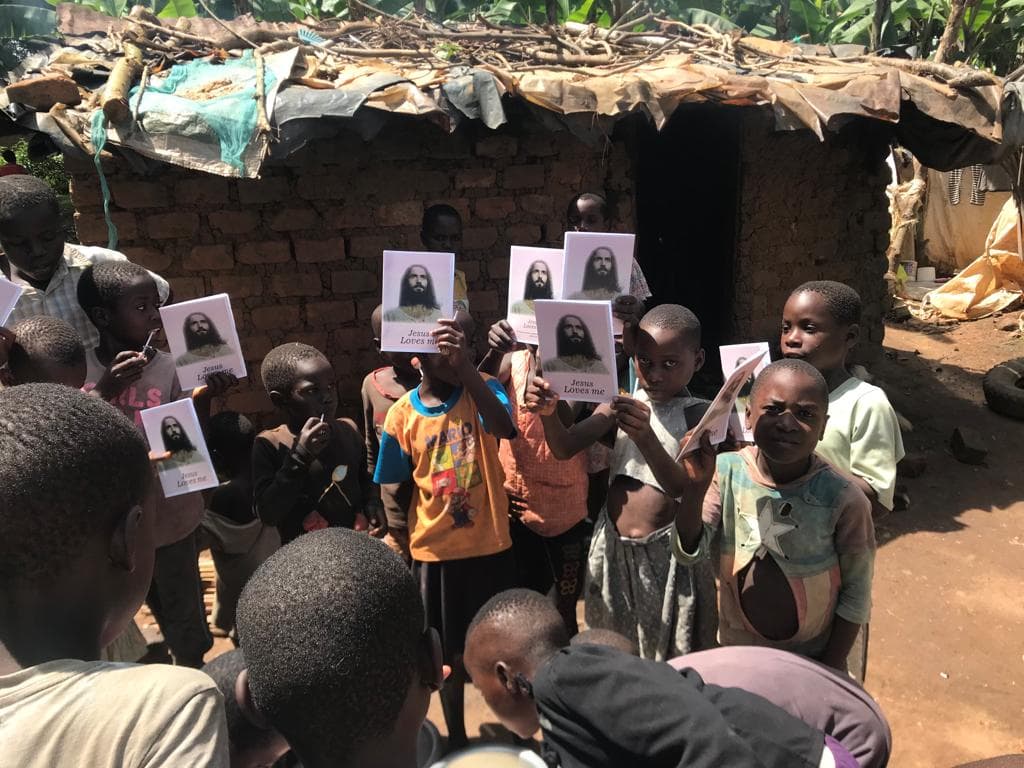 A group of children hold up "Jesus Loves Me" gospel tract photos outside a thatched hut.