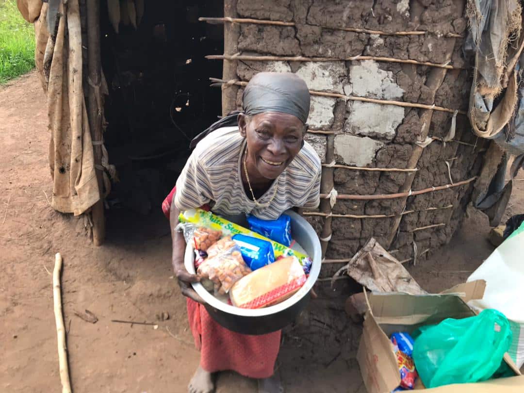 An elderly woman in a headscarf stands outside her mud-and-thatch home holding a basin of rice and sugar.