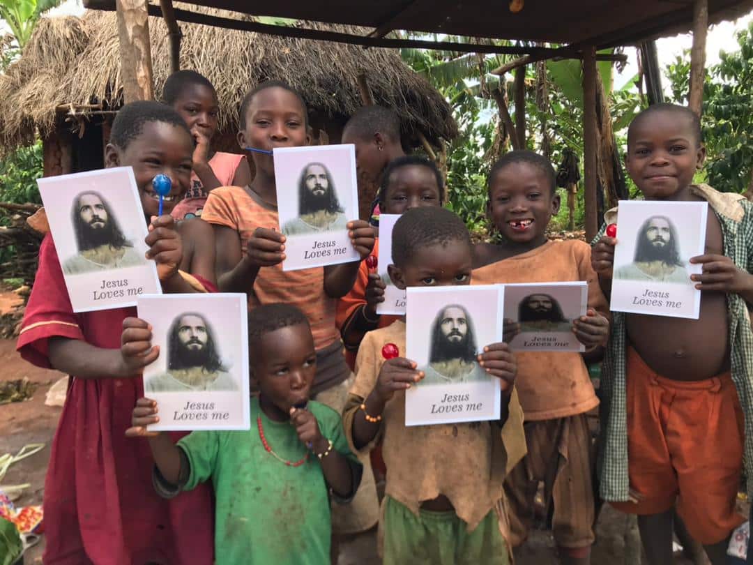 A group of children under a thatched shelter hold up "Jesus Loves Me" gospel tract photos.