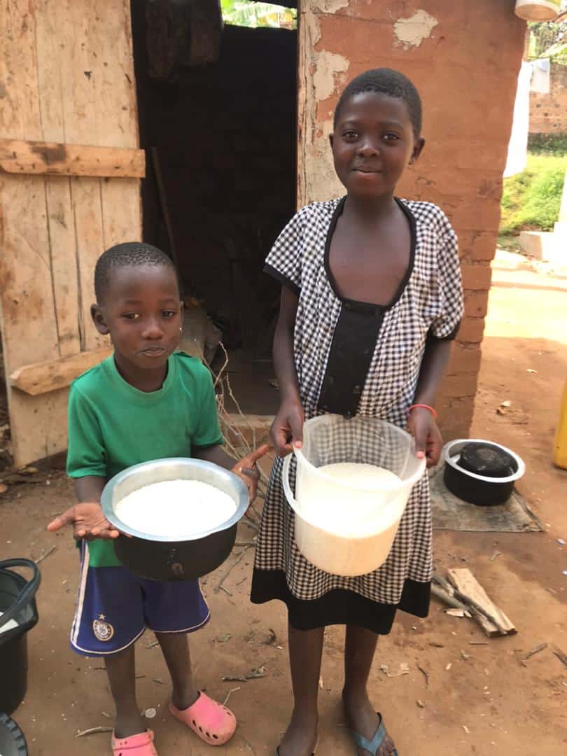 A woman and a young boy stand outside a home holding empty metal bowls, waiting for food.