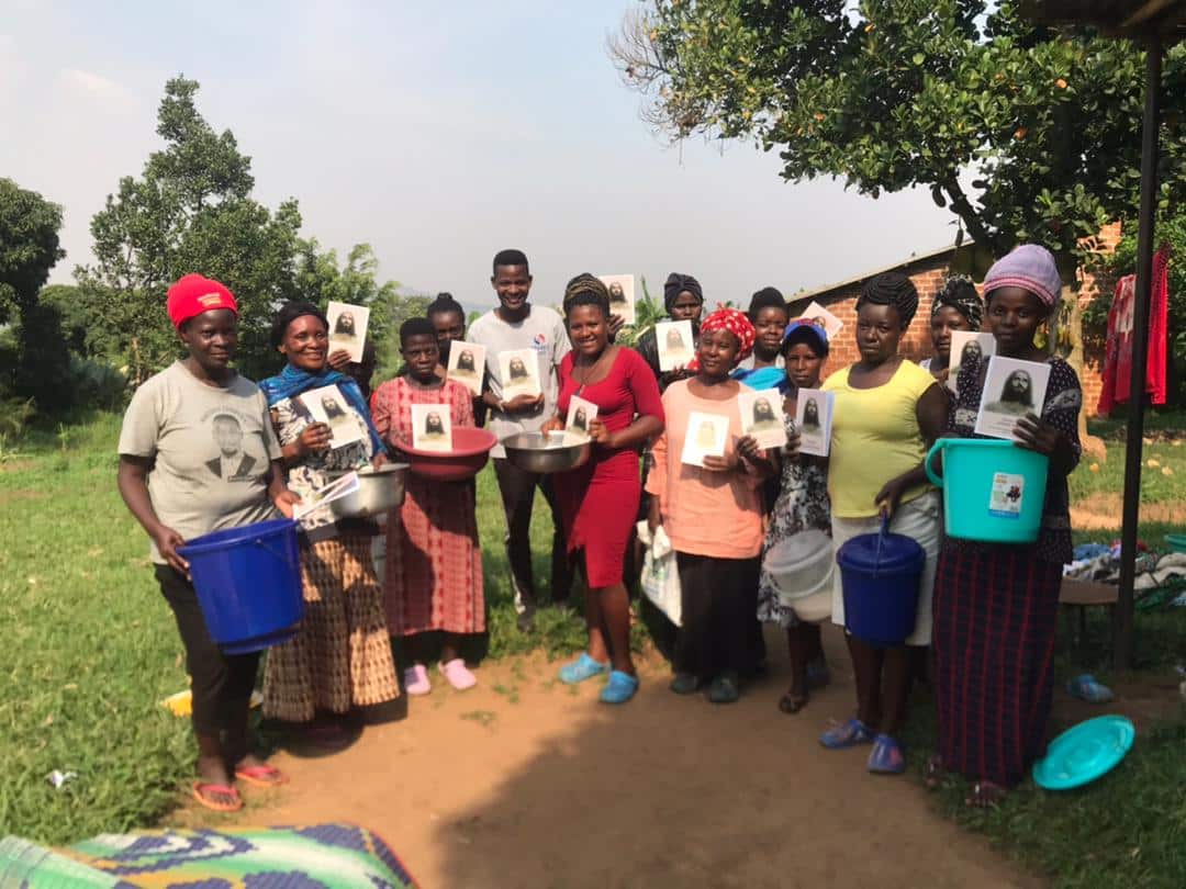 A line of women stand outdoors holding blue basins and gospel tract photos while receiving food gifts.
