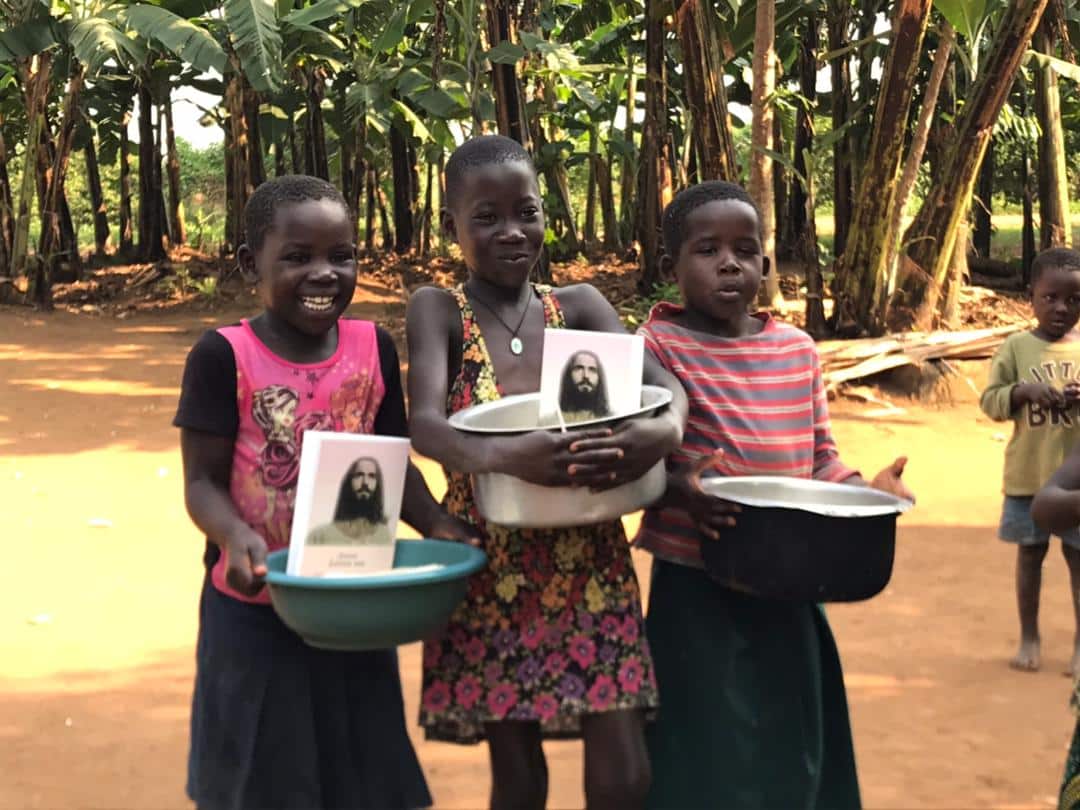 A group of children stand outdoors holding basins and a gospel tract photo near a banana grove.