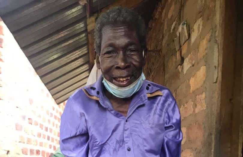 Elderly man in a purple shirt and face mask smiles while standing on a covered porch.
