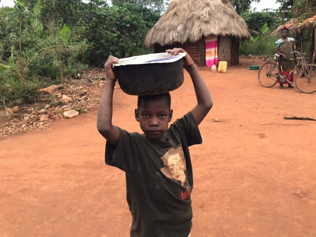 A boy carries a black basin on his head while walking down a dirt path past thatched huts.