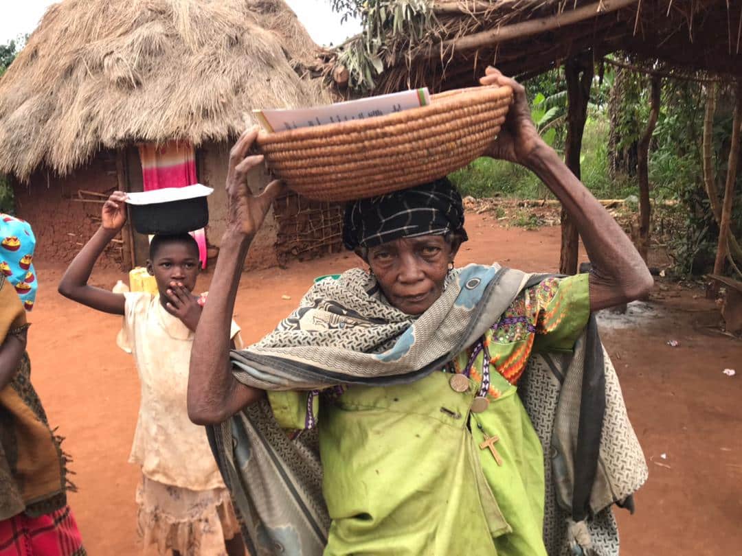 Elderly woman balances a basket and metal bowl of food on her head outside a thatched-roof hut in Mabu village.