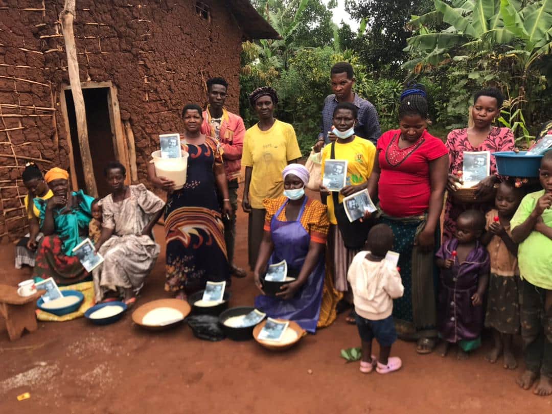 A group of women and children stand together outside a mud-brick home with bowls of food set on the ground before them.