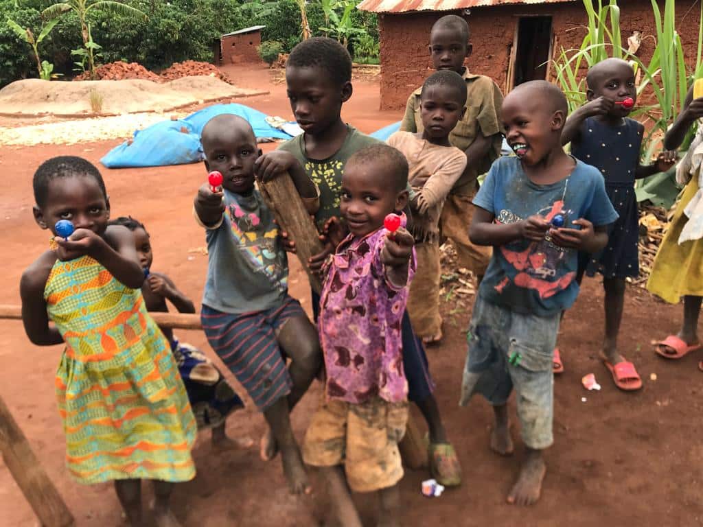 A group of young children stand together outdoors near a mud hut and banana plants.