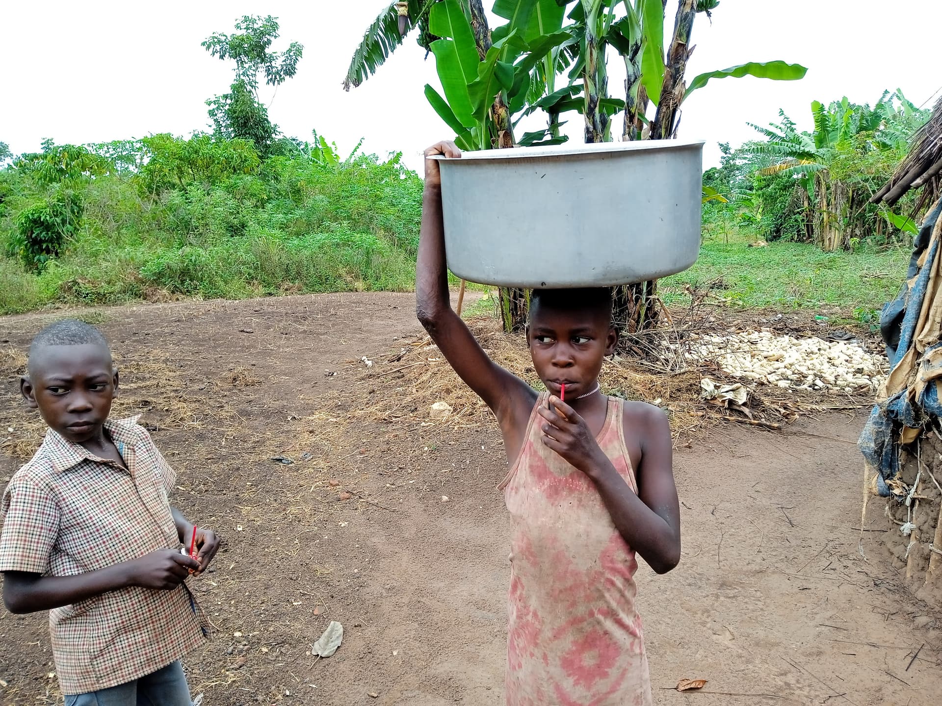 A girl carries a large white metal basin balanced on her head while walking on a dirt path, another child watching.