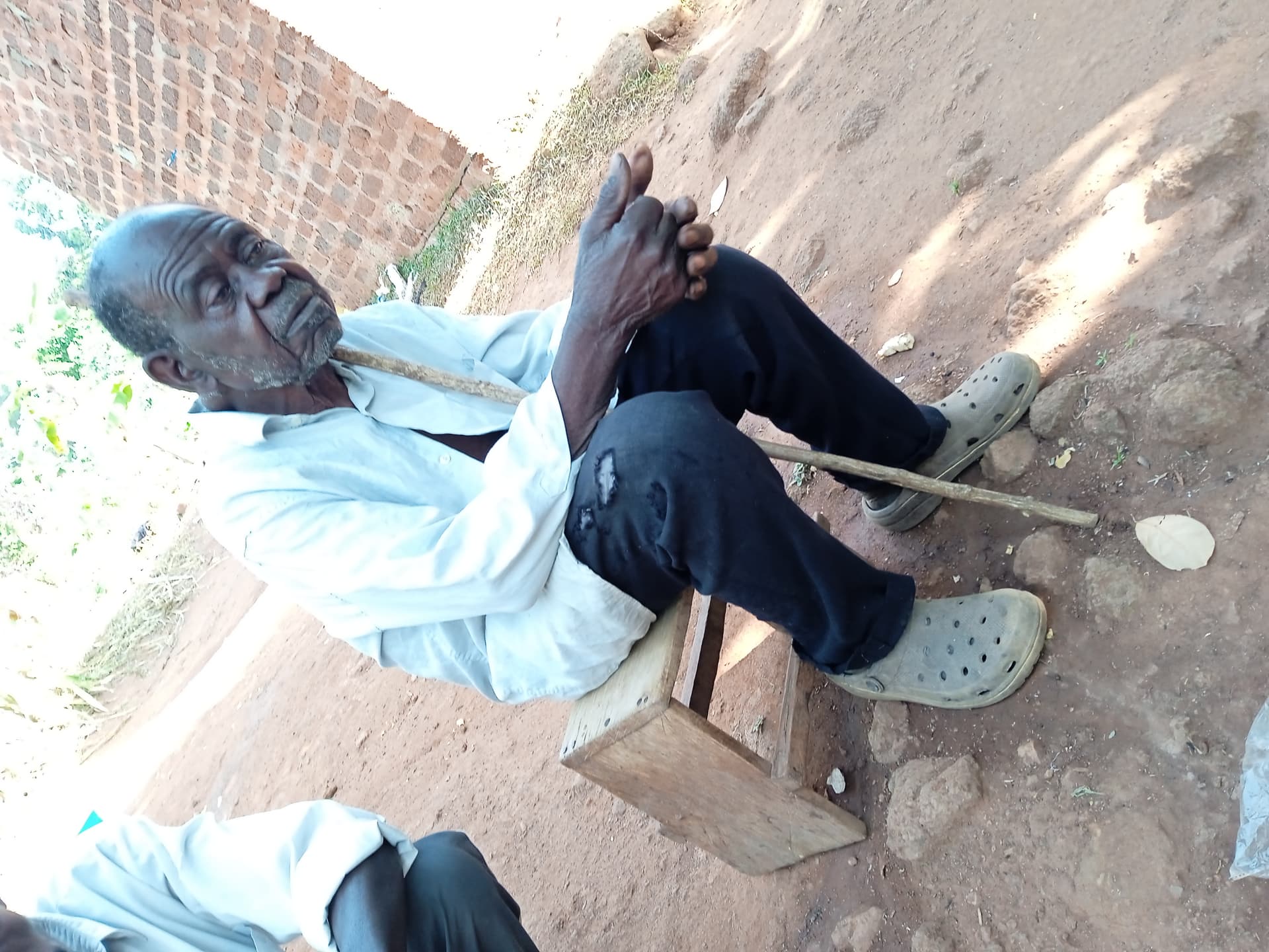 An elderly man sits alone on a wooden bench outside a brick building, resting on a cane.