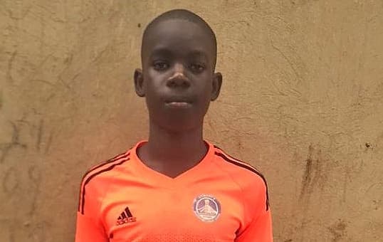 Joseph, a teenage boy in an orange soccer jersey, poses for a portrait against a mud brick wall.