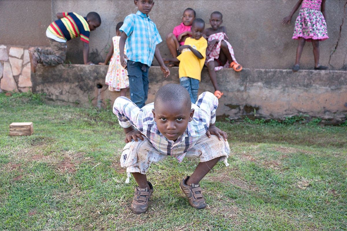 A young child in a patterned outfit crouches on grass in a yard, other children visible near a low wall behind.