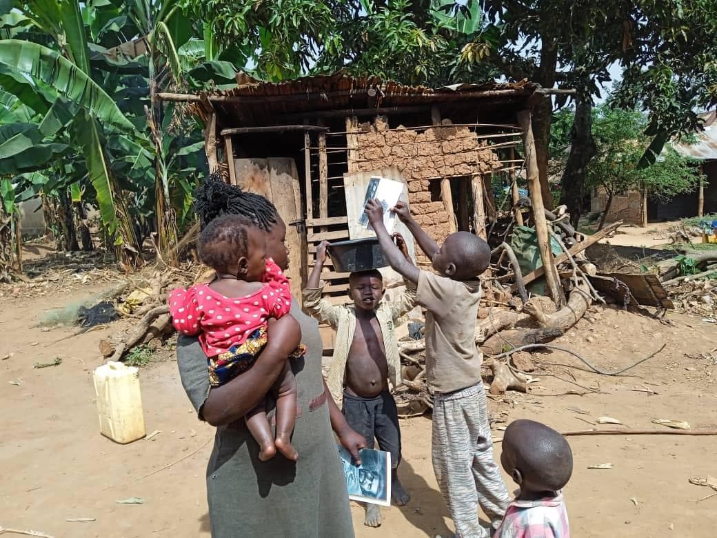 A mother carries a baby in a pink sling while a boy beside her holds up a white food container outside a mud hut.