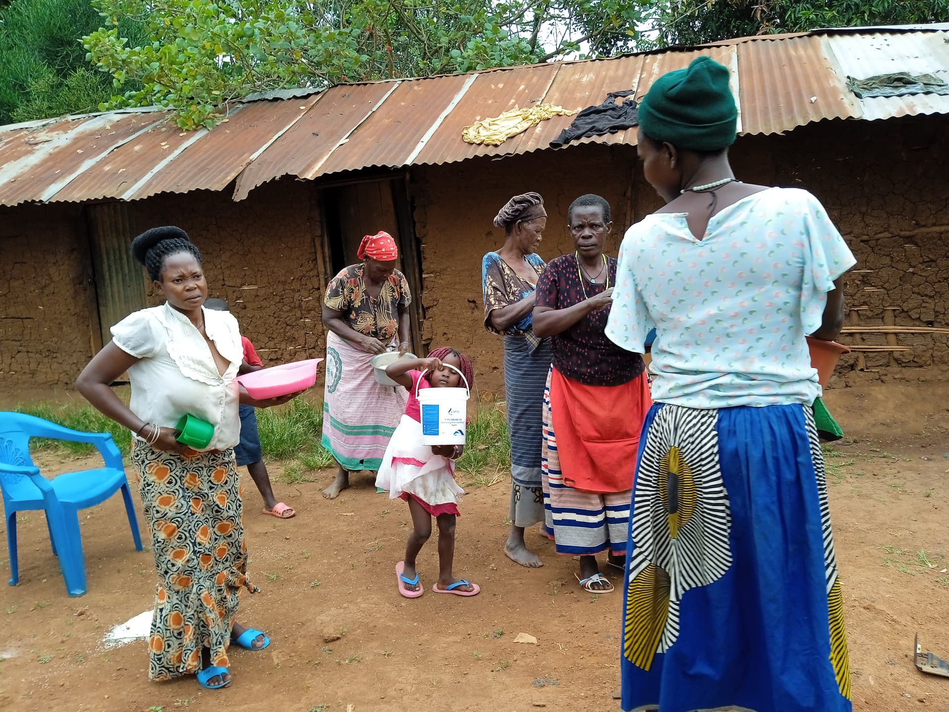 Women and children gather outside a thatched-roof mud home during a community visit, one child in a blue skirt nearby.