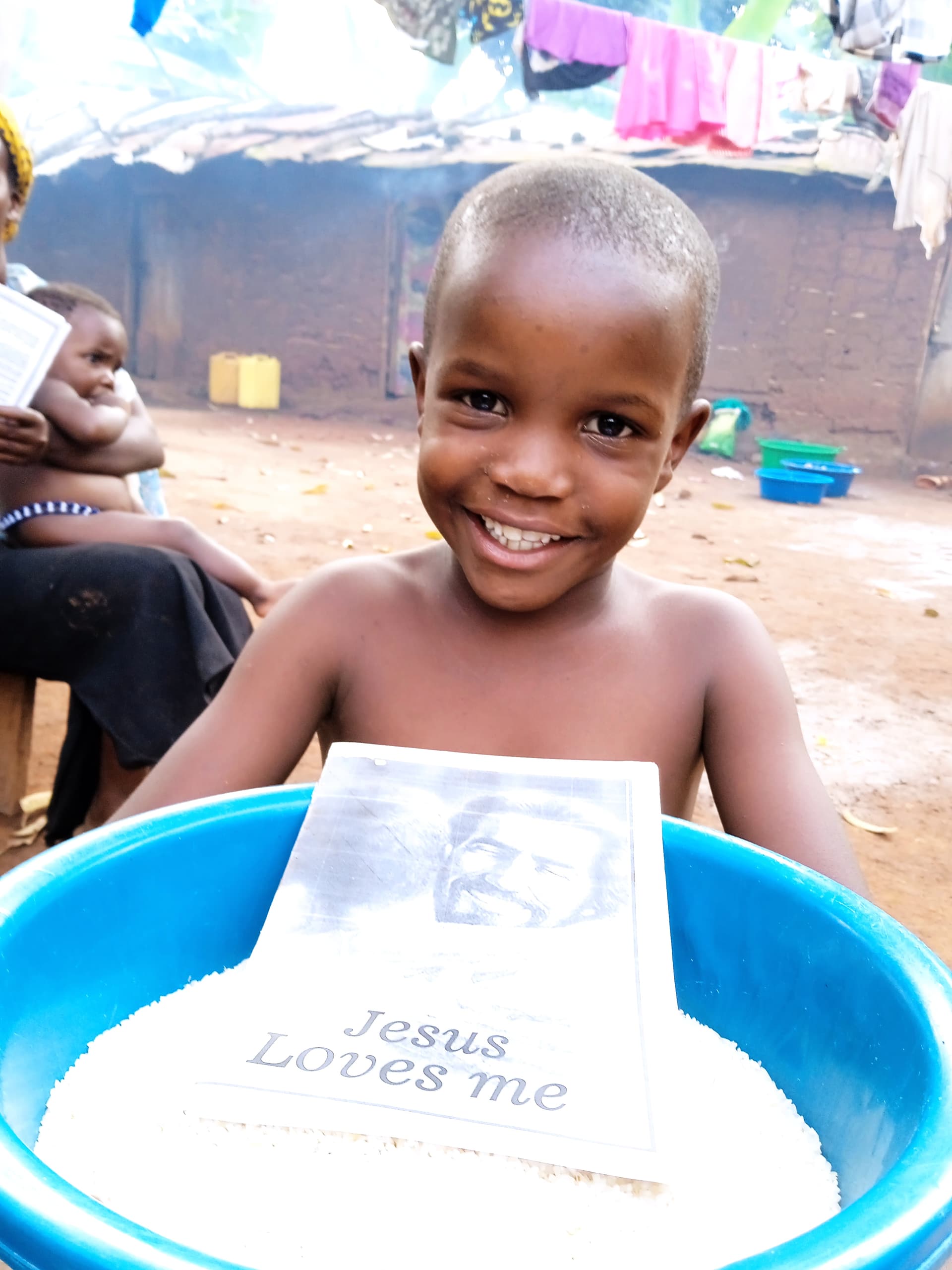 A smiling child stands behind a blue basin holding a Jesus Loves Me tract, another person visible in the background.