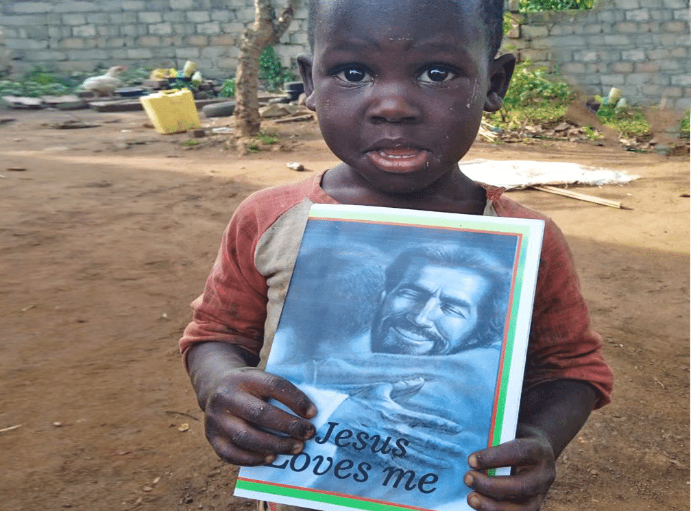 A young boy stands outdoors holding a Jesus Loves Me gospel tract in front of a brick wall.