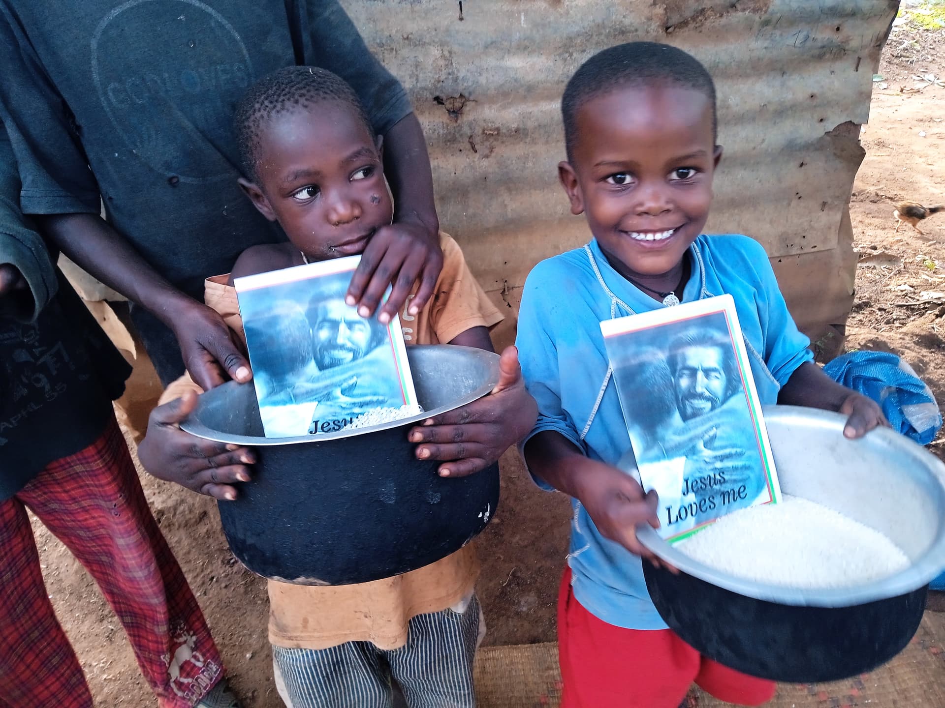 Two smiling boys hold bowls of rice with Jesus Loves Me gospel cards tucked inside, standing outdoors.