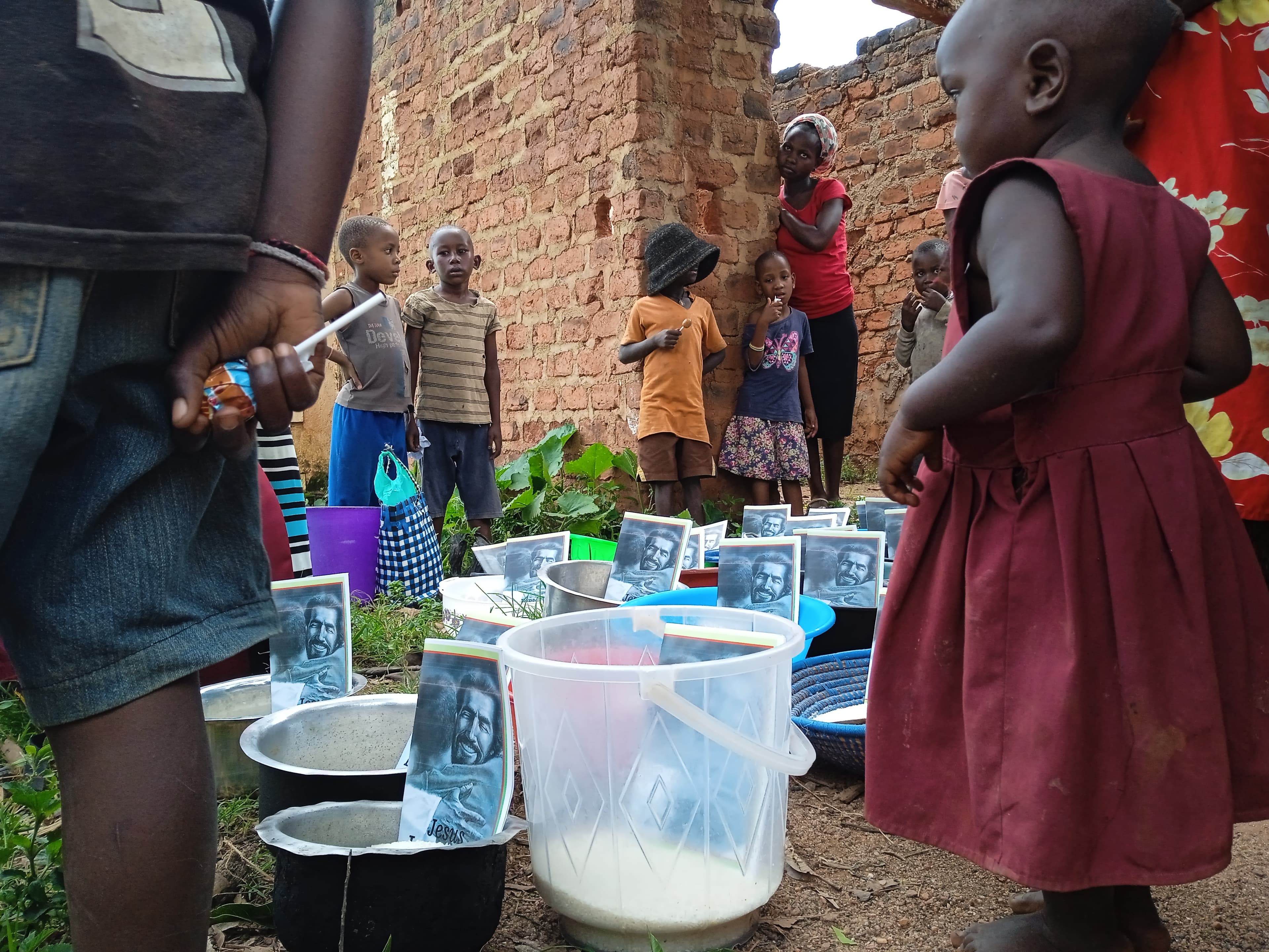 A child in a maroon dress stands in a line of children near a brick wall, holding a cup over a black bucket of rice.