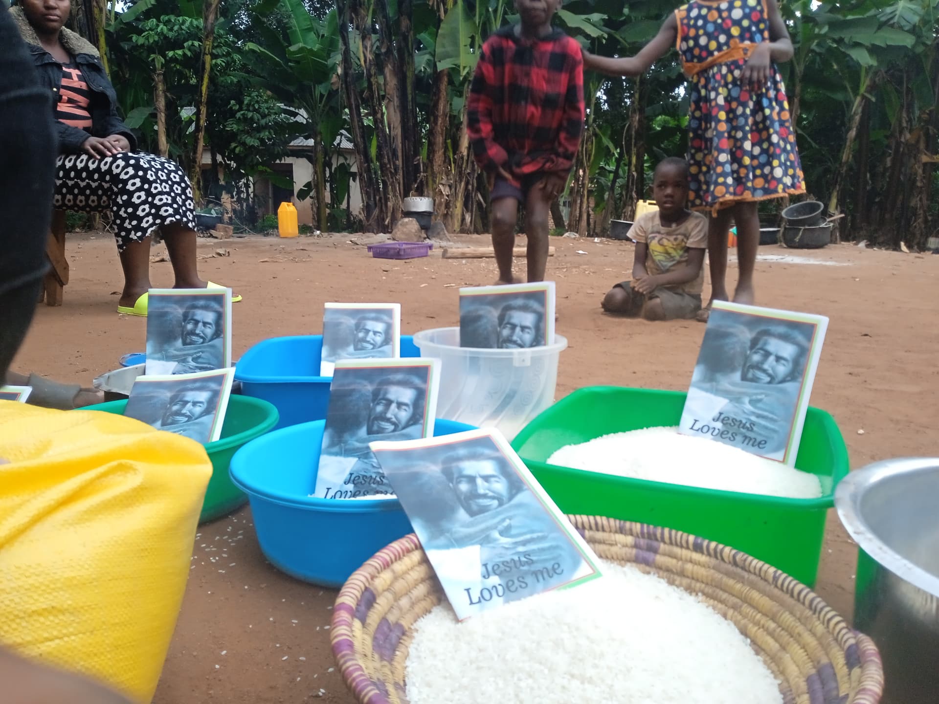 Women and a girl in colorful dresses stand near green, yellow and white basins of rice with gospel tracts propped against them.