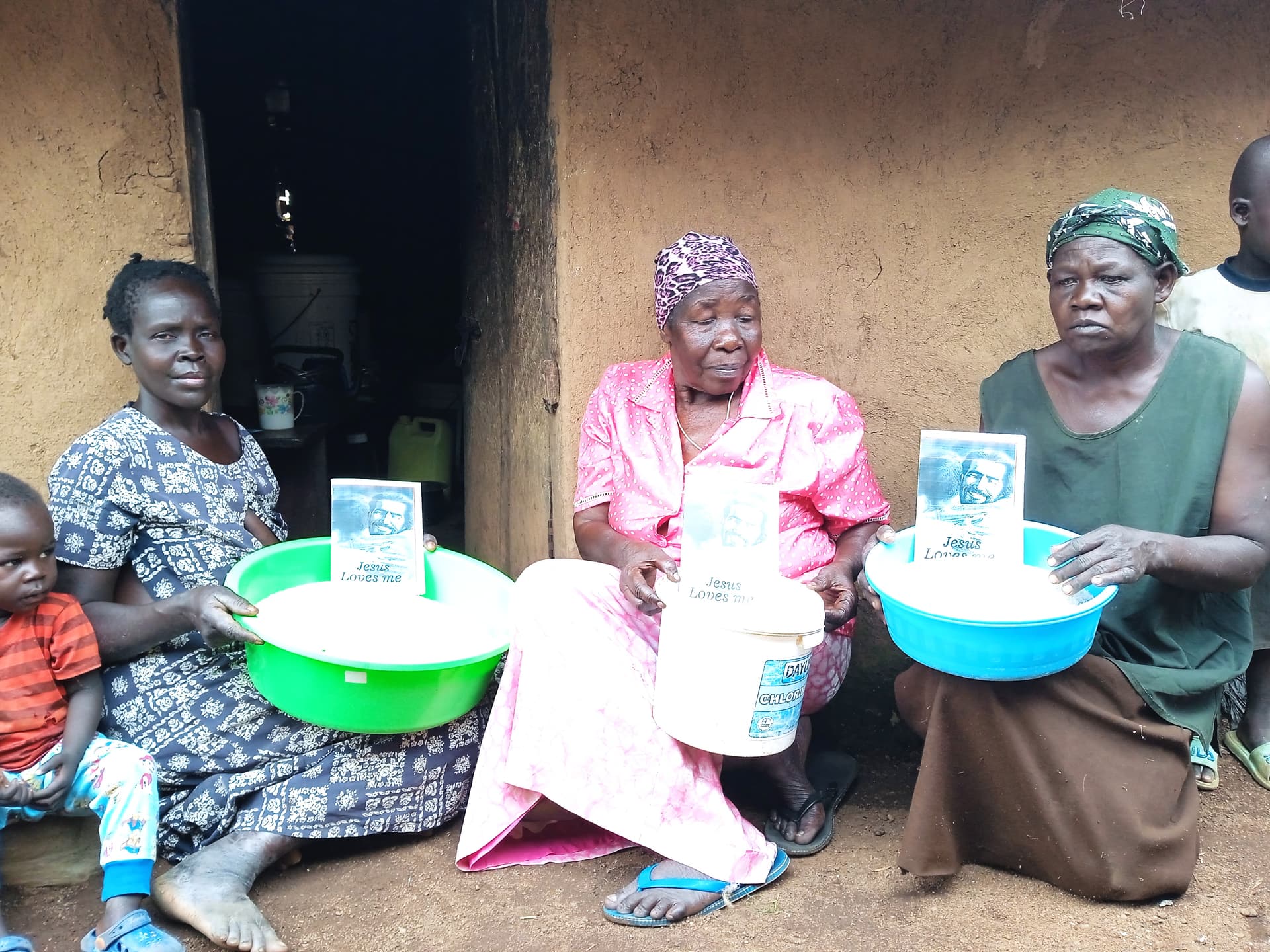 Two Ugandan women sit with a green and a white basin of rice, a JFM Jesus Loves Me card propped against one basin.