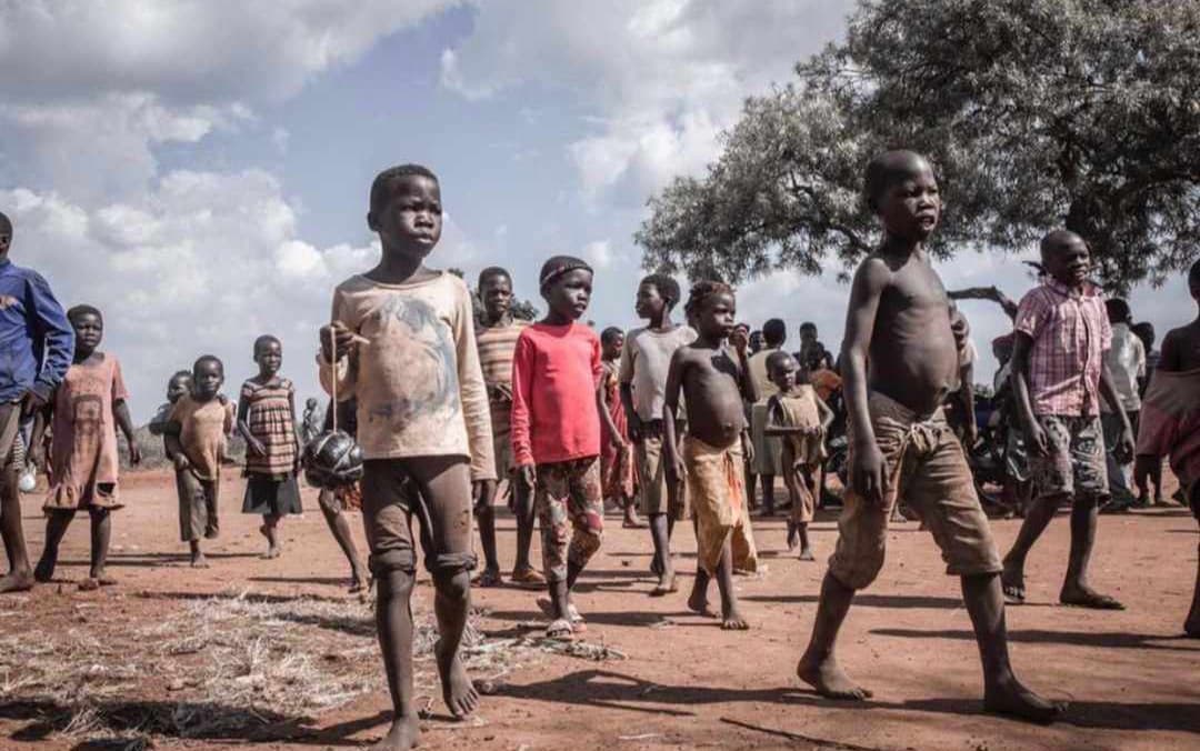 A group of barefoot children and young men stand and walk across an open dirt field under a cloudy sky.