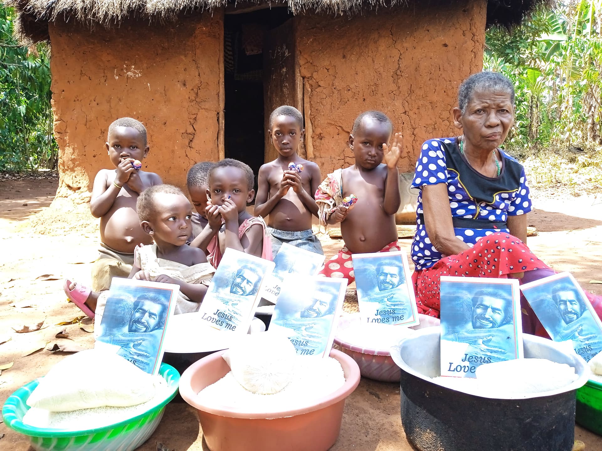 An elderly woman sits among a group of children outside a mud hut with food basins nearby.